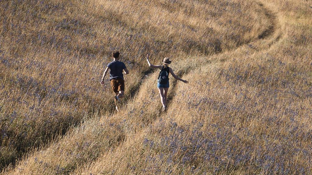 Luke & Nell walking along a narrow winding path through a golden meadow up on Monte Subasio near Assisi in Umbria.