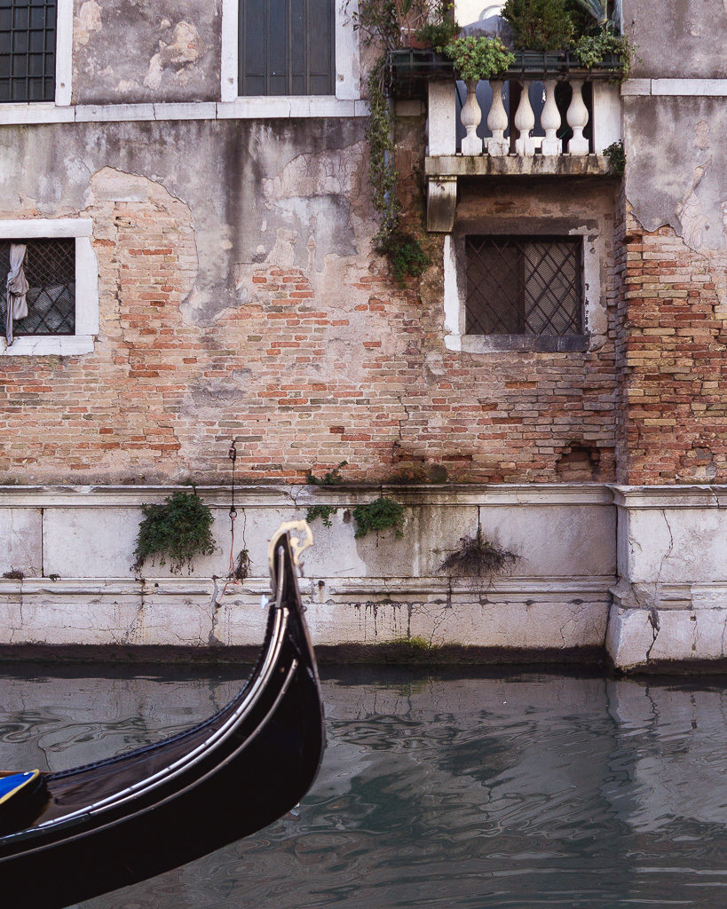 Gondola in bottom left corner of photo with worn Venetian building in background across quiet canal