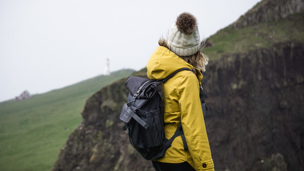 Nell showing what to pack for hiking in the Faroe Islands. Always carry a hat and a warm, waterproof coat. Shot from behind Nell, quite close, and she is looking into the distance at the Mykines lighthouse.