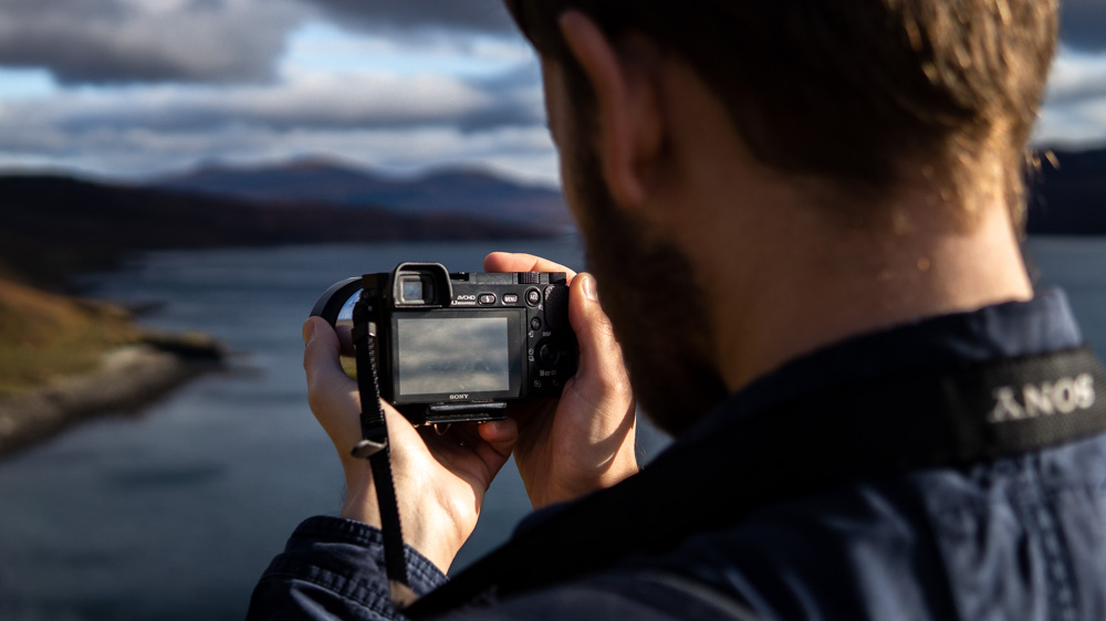 Image taken over Luke's shoulder showing him holding a Sony a6000, looking at the screen on the back, checking the shot which is of the coastline of the Isle of Skye.